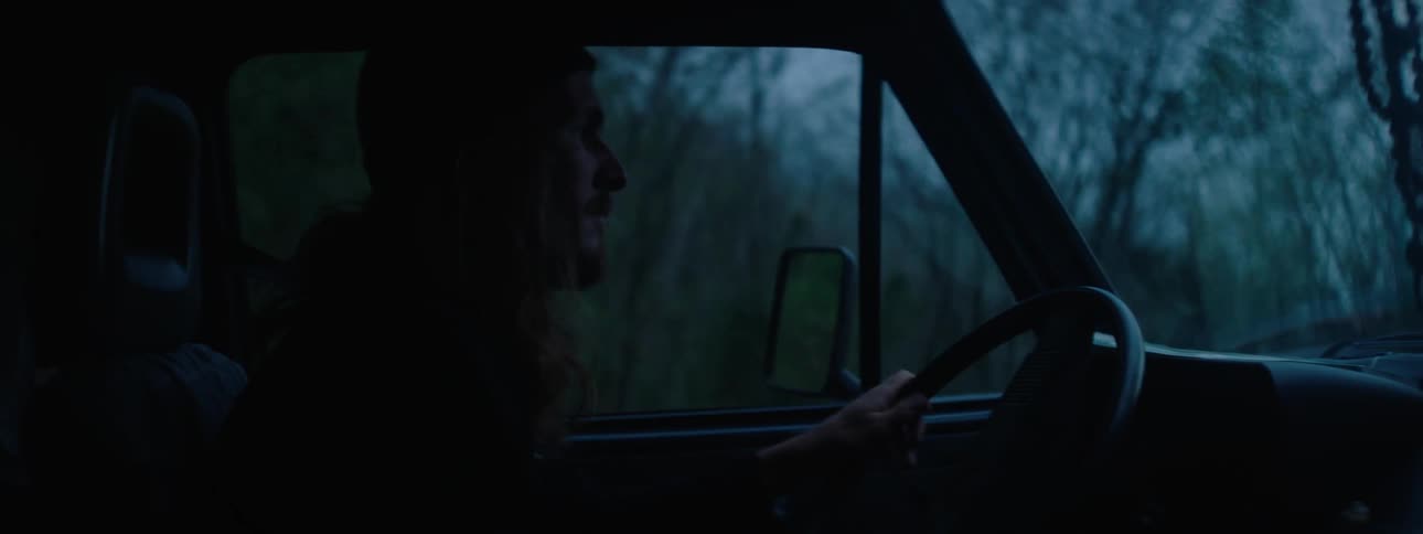A long haired man is drivingan automobile through a rural area and he looks out his side window as he drives