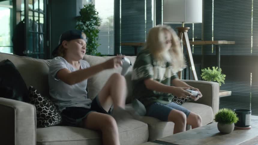 Two young kids brother and sister arrive home from school jump on the couch and begin to play video games with playstation controllers