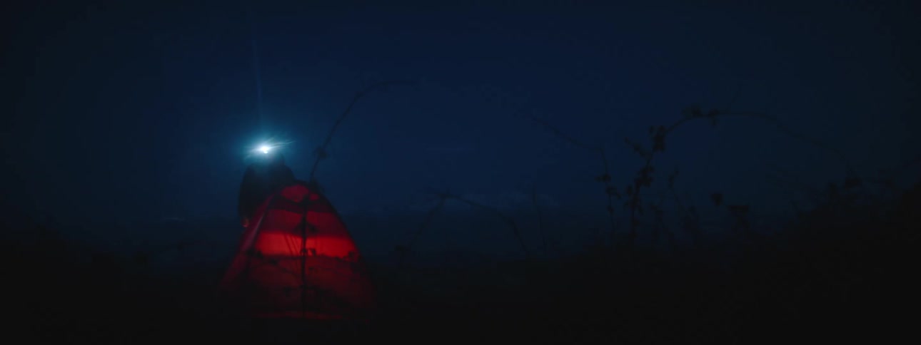 A man in profile at night wears a headlamp and moves from crouching to standing by a tent outside