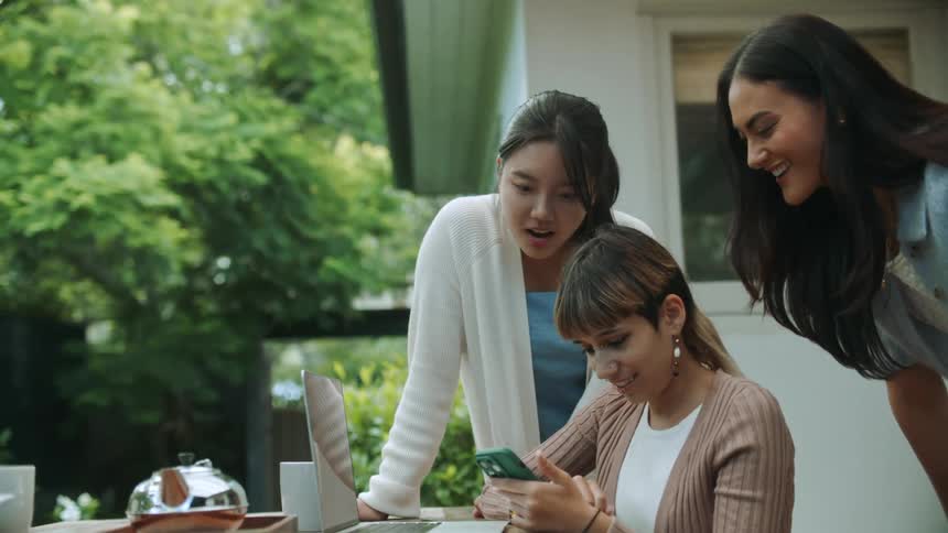 Two women standing outside leaning over a third women sitting down as they all look at a cellphone