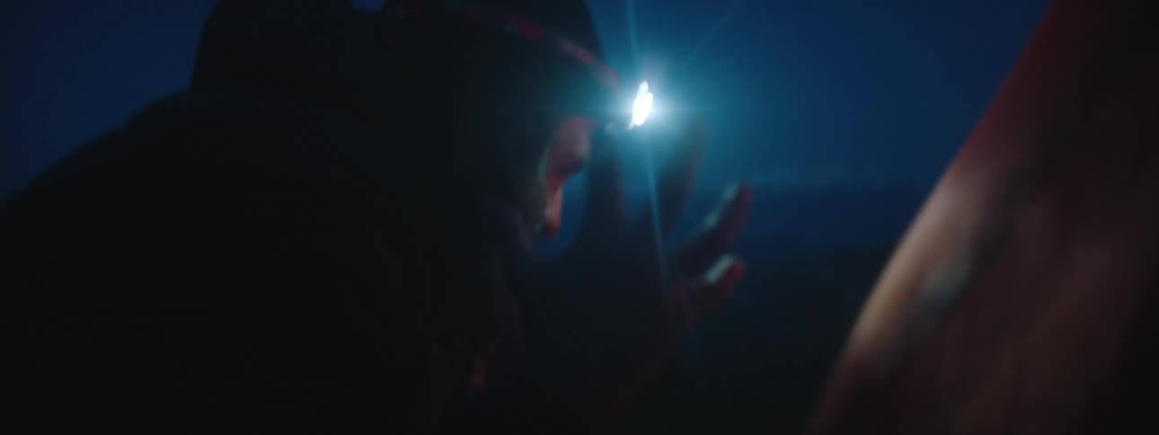 A man wearing a head lamp setting up a tent in the blowing wind at night