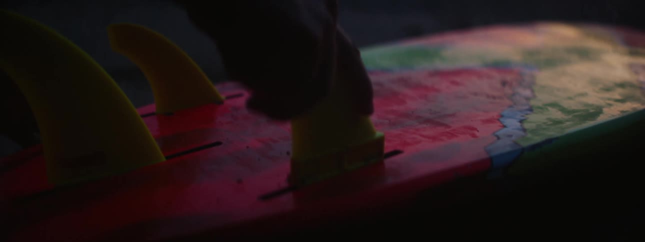 Male surfer partially silhouetted against red sky attaches fins to surfboard lying on beach