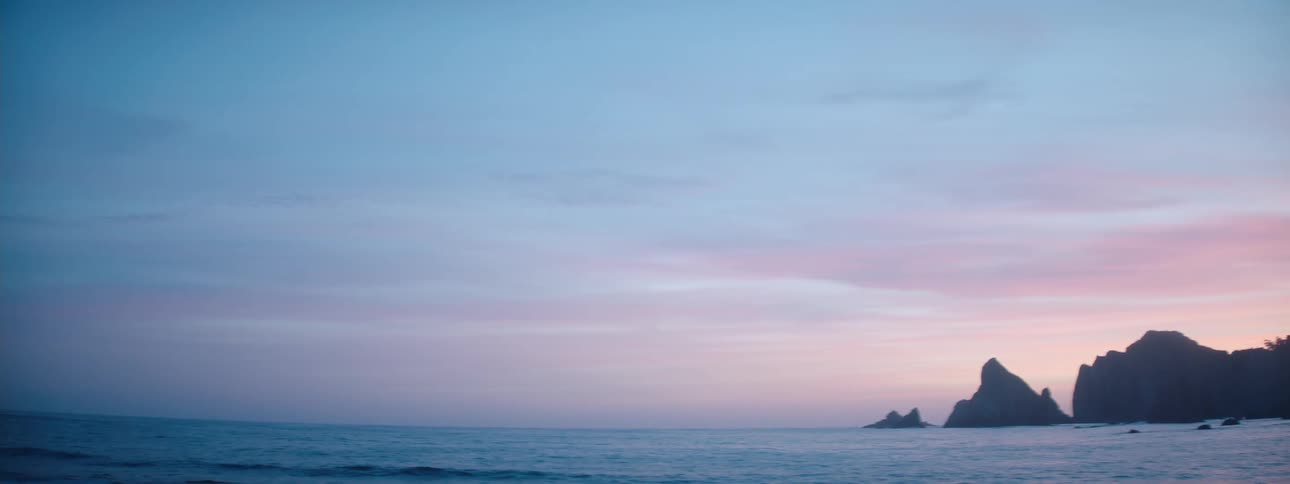 A wide shot of a pink and blue dusk sky over an ocean and coastline cliffs