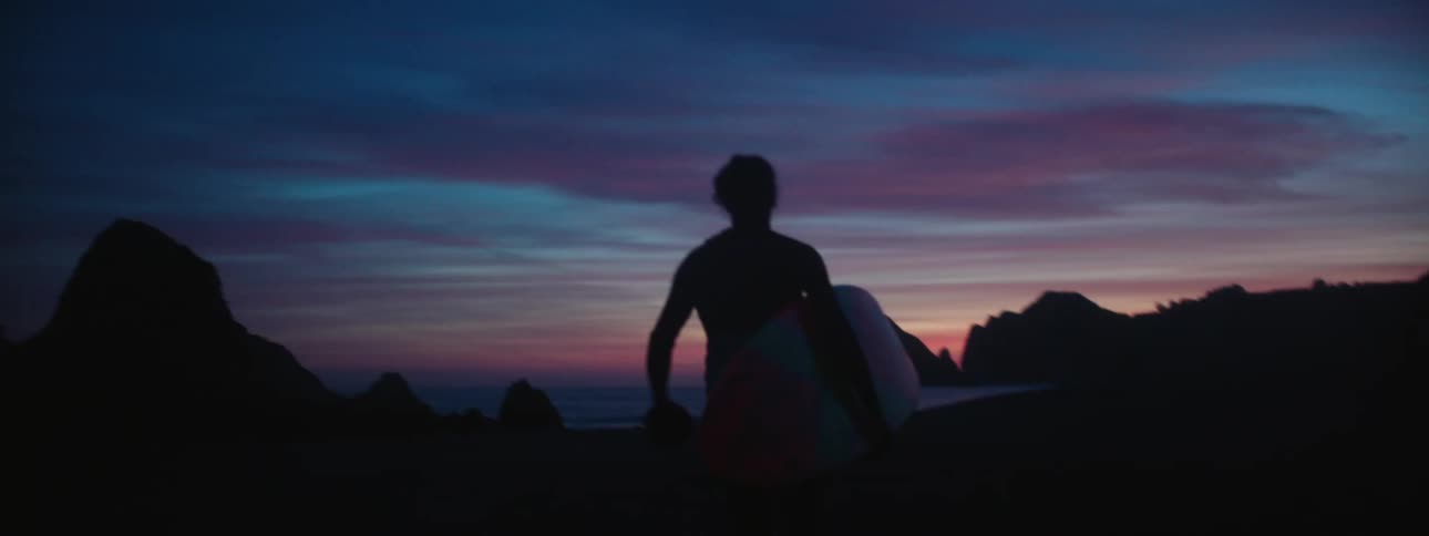 A man is walking on the beach with a surf board at dusk