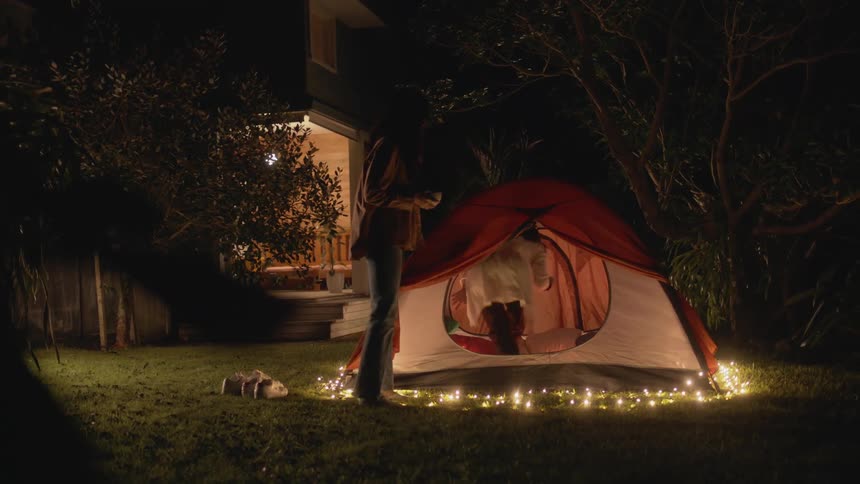 Two teenage girls are carrying bowls through the yard and into a tent at night