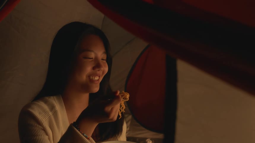 A woman sitting in a tent smiling and laughing while eating noodles out of a bowl