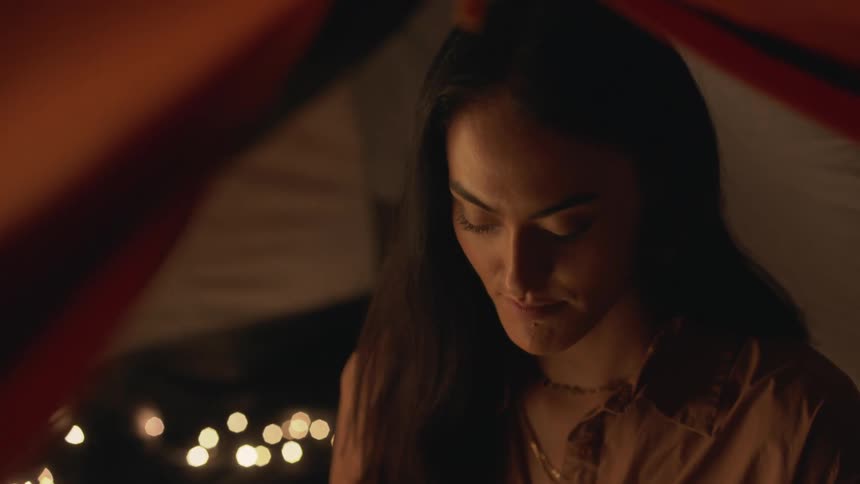 A woman with long dark hair smiles as she is talking and looking down at something in front of her in a dark place with lights reflecting behind her