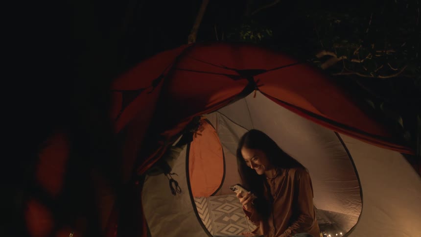 A woman is sitting inside a tent holding a playing cards in her hand as she holds her phone in the other hand she smiles and laughs as she talks