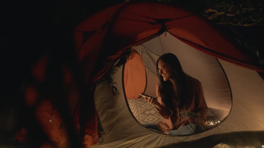 A teenage girl with long dark hair is in a tent smiling and laughing as she talks on her phone