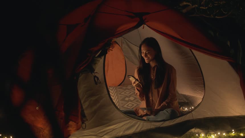 A woman with long dark hair is sitting inside a tent holding playing cards in her hand as she holds her phone in the other hand she smiles and laughs as she talks