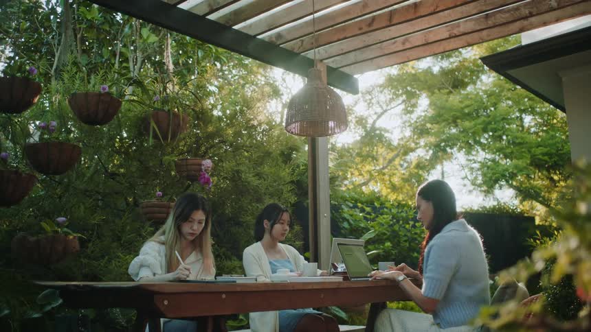 Women sitting at table use laptops and write in notebook in backyard