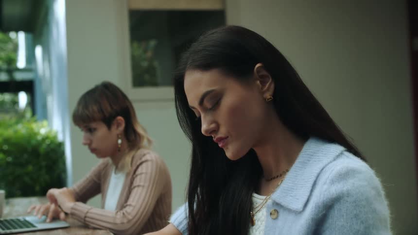 Two women sitting outside at a table one using her laptop and the other writing