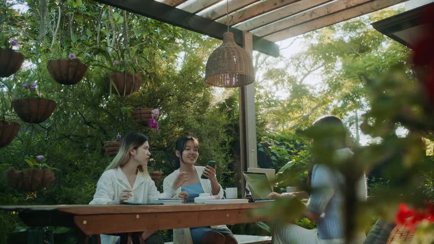 Three teenage girls are sitting at a table outside as one girl types on a laptop computer and another girl is showing them her phone