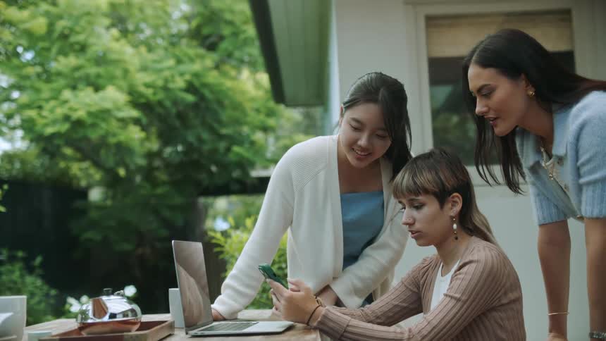 Two women standing over a third woman sitting down while looking at the phone in her hand