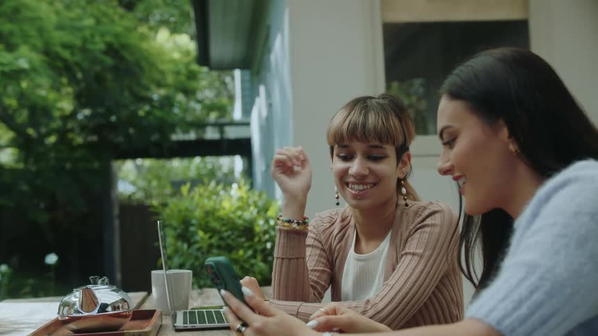 A couple friends sitting at a table in the backyard talk white looking at a phone