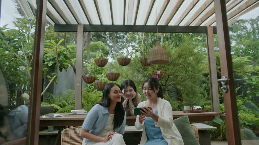 Group of friends sitting under a pergola in the garden laugh looking at a phone