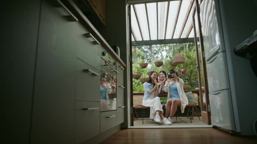 Three women sitting down outside under a roof posing for a photo taken by the girl with the phone