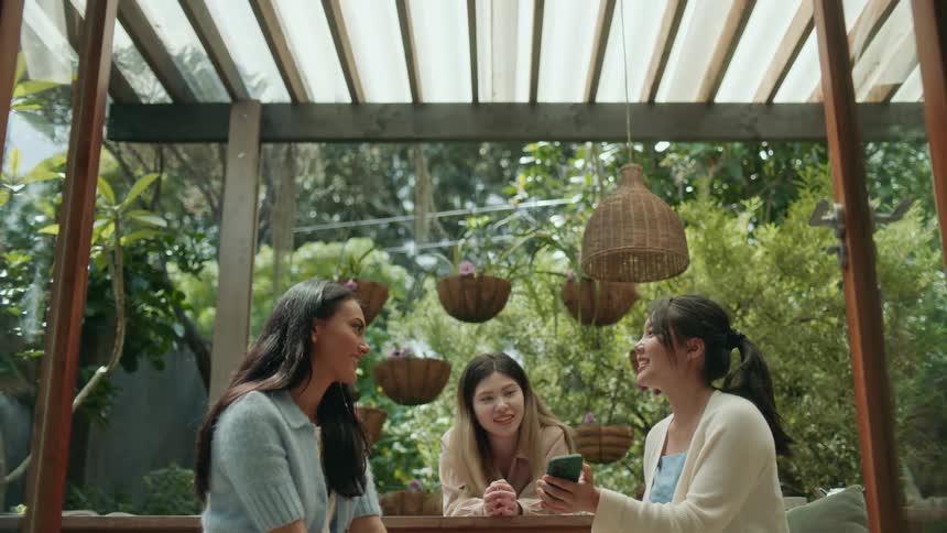 Three women sit around a table under a porch roof and look at a womans phone she has in her hands as they all laugh and smile