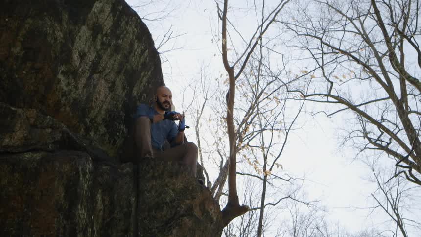 A man sits on a rock ledge and adjusts his gloves on a sunny day