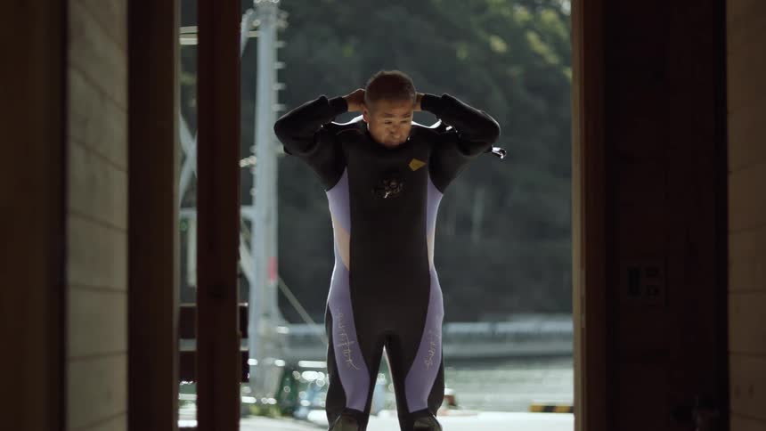 A man stands in a doorway putting a wetsuit on and then turning around and walking away
