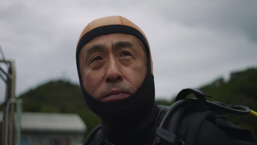 Man wearing wetsuit stands on boat with forest covered mountains in background