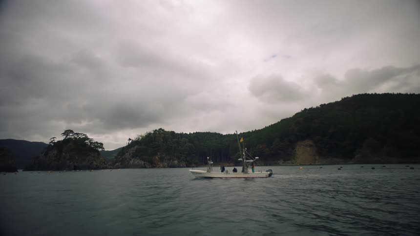 Boat moves through water past buoys and floats with forest covered hills on shore in the background