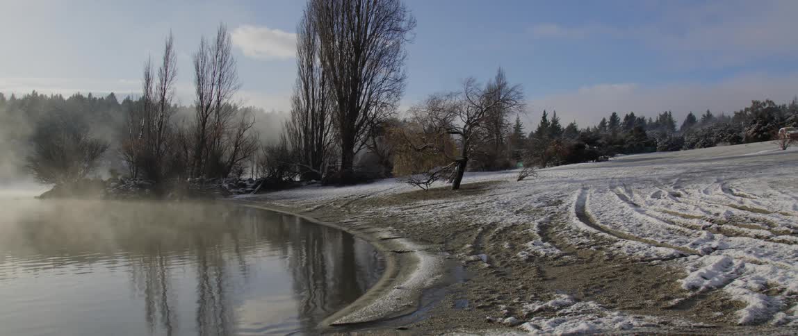 Mist or fog over the calm surface of a lake next to a field covered in snow at morning