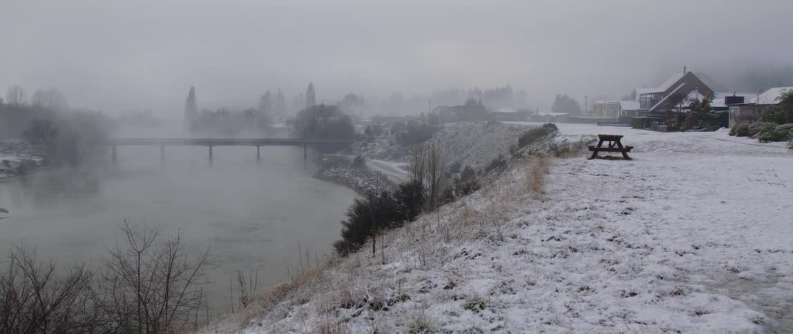 Snow covered ground and fog settling over a rive and bridge showing a village in the distance