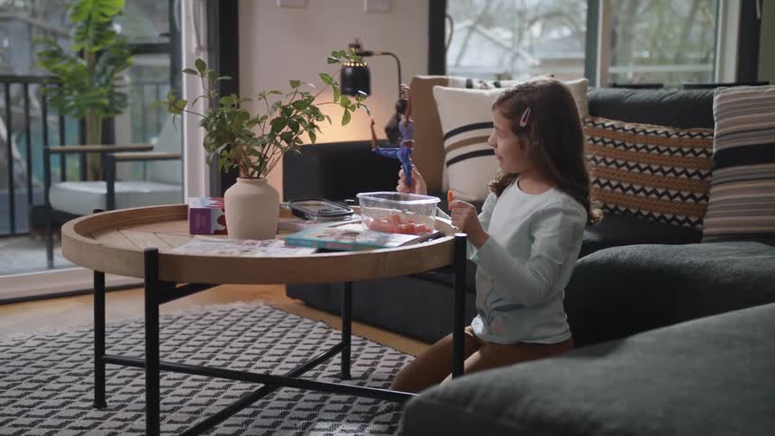 A young girl eating a snack and playing with a barbie doll at home in a living room