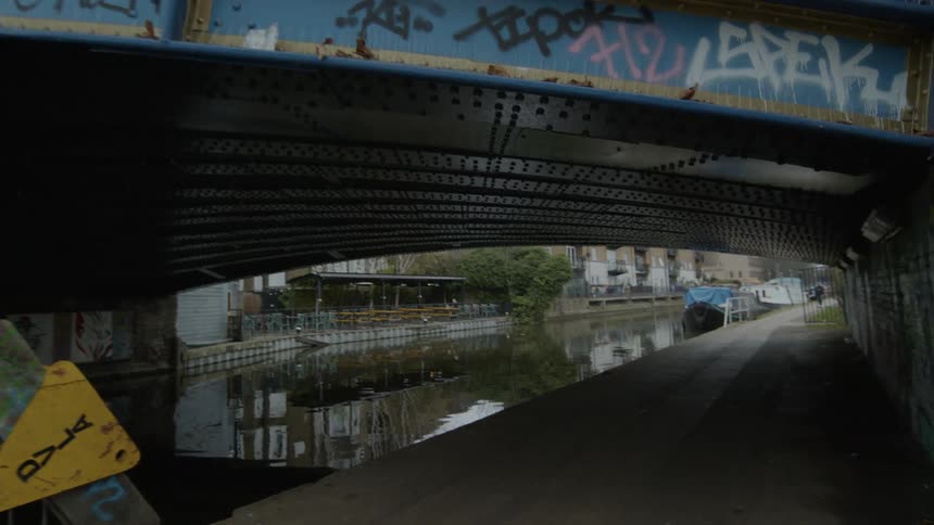 Two people are walking on a bridge as a train passes and the water underneath sparkles in the sunlight as an apartment building is seen through the tunnel