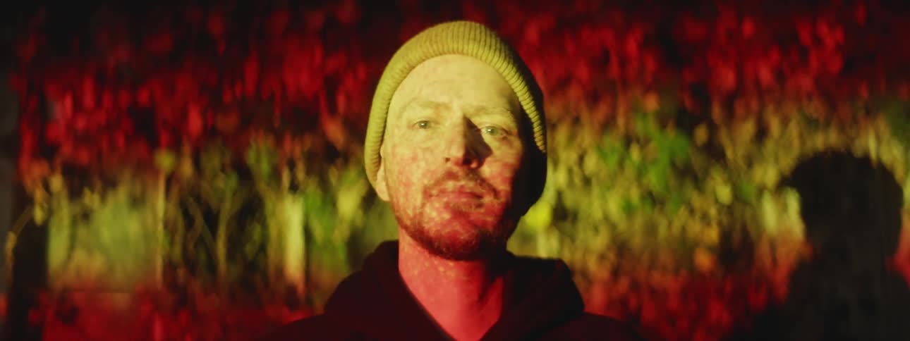 A man is wearing a knit hat and coat as colored lights move across his face and on the wall behind him