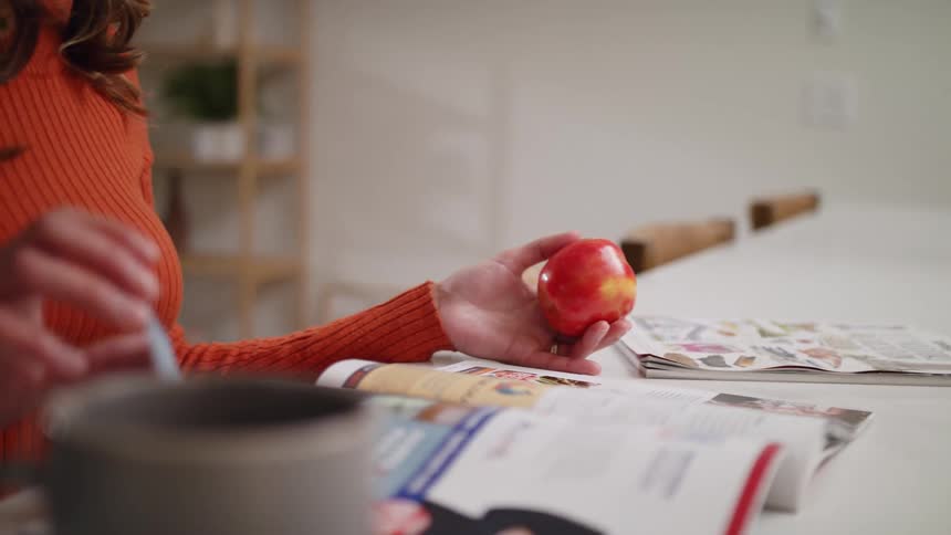 Woman with brown wavy hair and red sweater leafs through some magazine holding a red apple