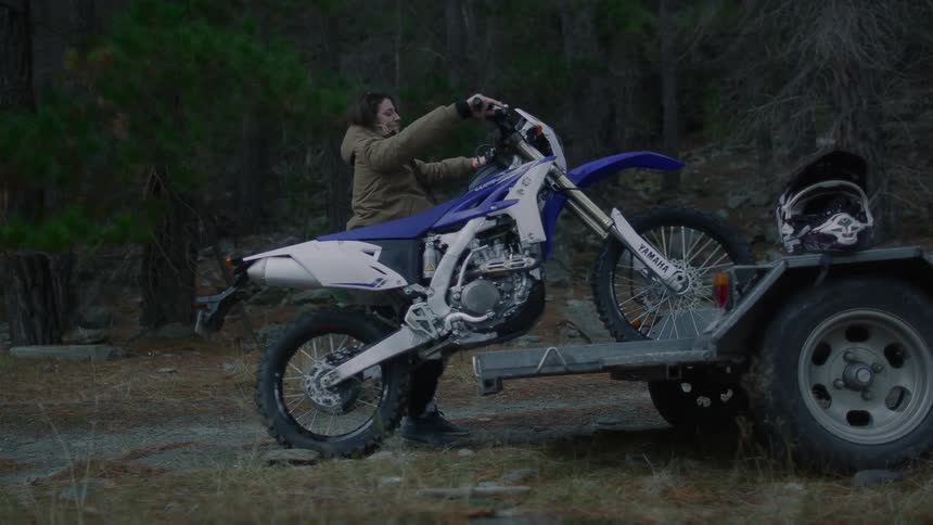 A woman unloads an off road motorcycle from a trailer in a woodland during the day