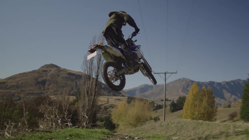 A motorcycle travels over a hill and leaps into the air and flies before landing back on the ground and there are mountains in the distance