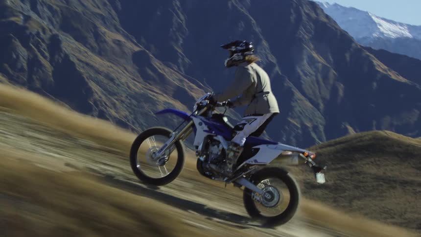 Motorcyclist stands while riding motorcycle up slope overlooking valley with mountains in the background