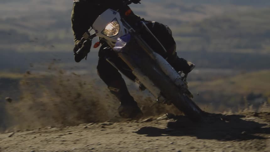 A person wearing a black hoodie and helmet riding a dirt bike on a dirt track going around a corner kicking up dirt and dust