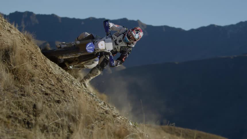 A person riding a dirt bike up a hill and skidding around a corner and back down the hill with mountains in the far distance
