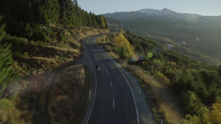 Motorcyclist speeds along highway hugging side of forest covered mountain over valley