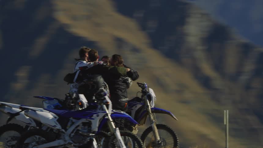 A small group of people are standing near their motorcycles in the mountains as they group hug and pat each other on the back