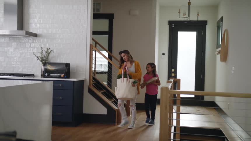 A girl follows her mother into the kitchen as she carries a bag of groceries
