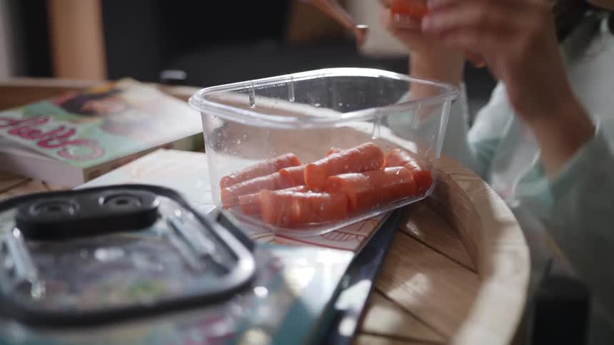 Girl holding piece of carrot plays with doll while sitting at table near container of carrots