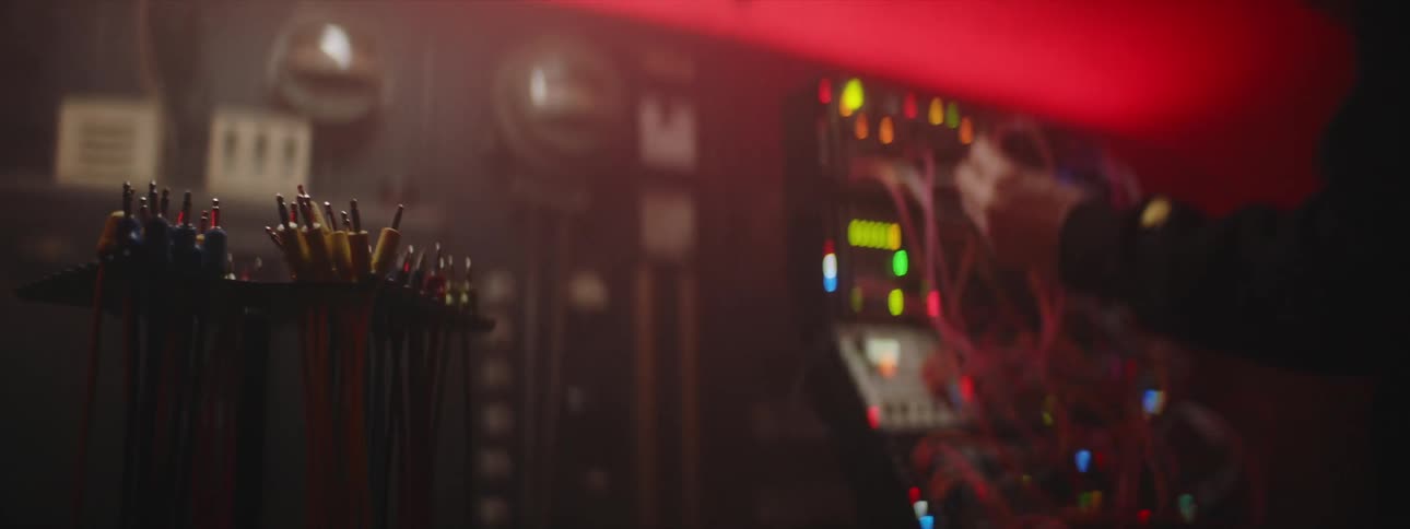 A man is standing in a smokey room as he takes a wire from a rack of wires and connects it to a computerized machine with many glowing lights
