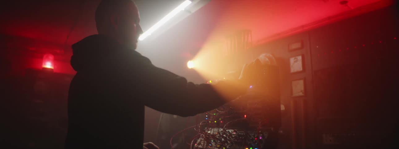 A man is standing in a smokey room in front of a computerized machine as he moves wires and colored lights flash and glow