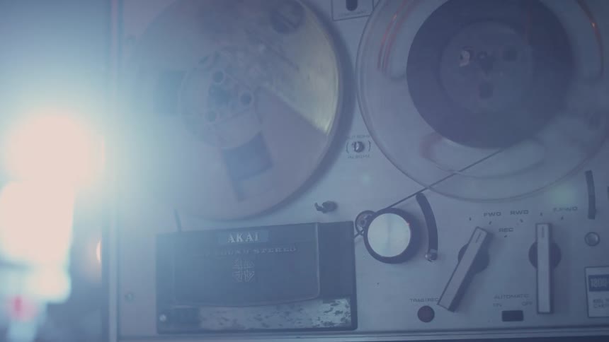 Two large film reels rotating around while tape is being fed into them and sunlight shines through the windows in the back