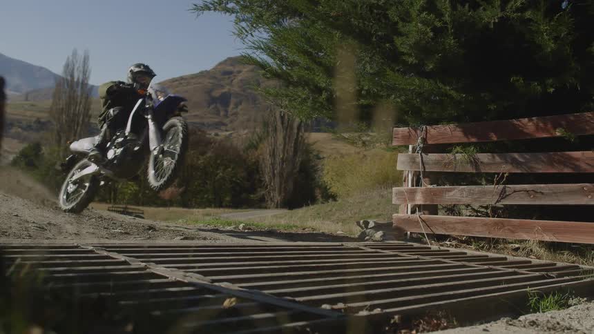 Motorcyclist crossing grate bridge over gulch pops a wheelie with mountains and highway in the background