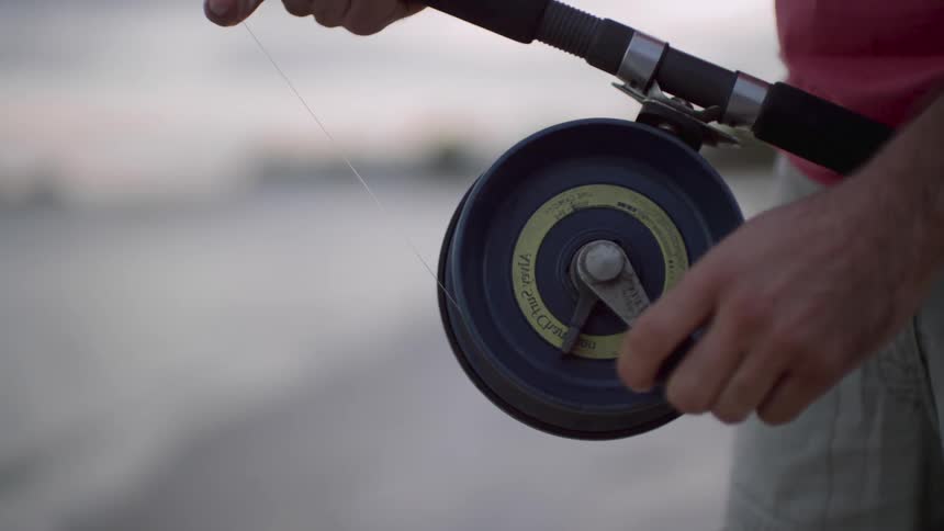 A man is standing outside holding a fishing rod and turning the crank on the reel