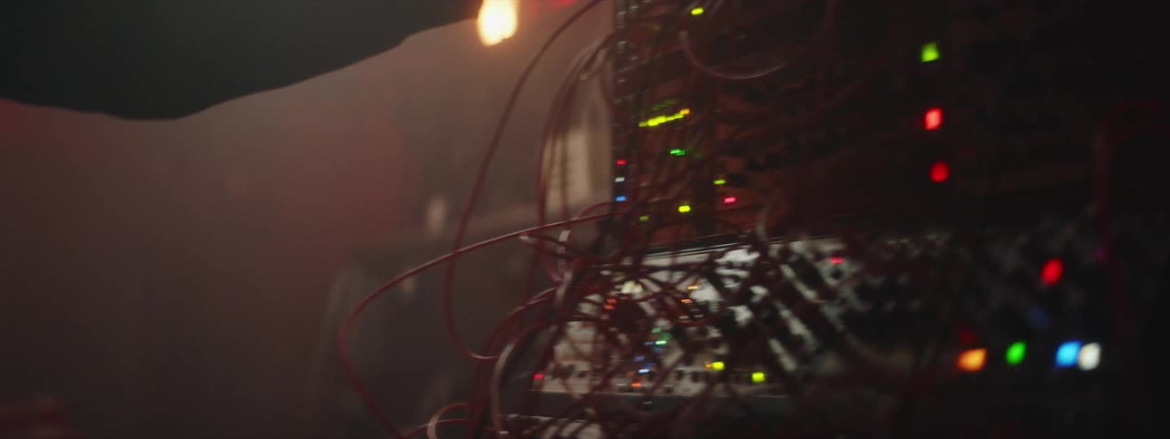 A man is standing in front of an electronic machine with colored lights as he moves wires and turns knobs