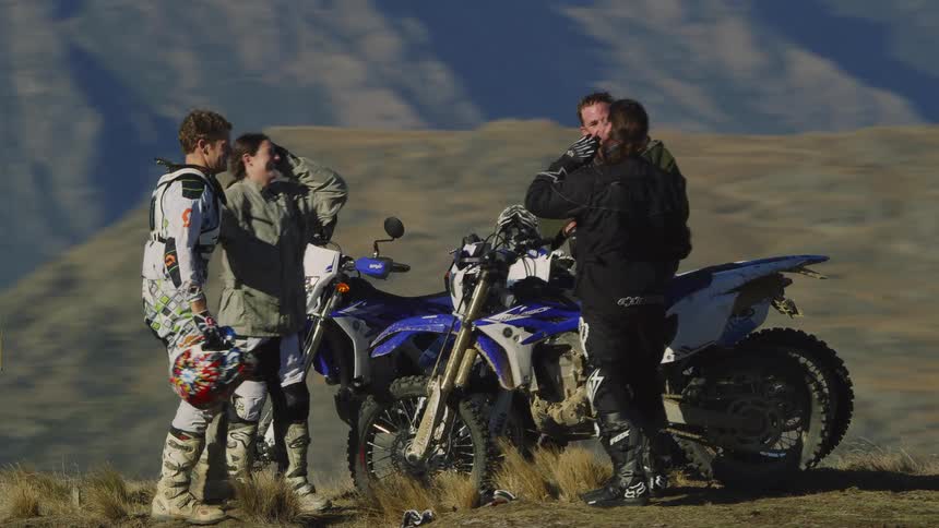 A small group of people standing near dirt bikes and hugging talking and smiling while they are on top of a peak