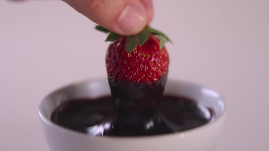 A person is handdipping a strawberry into a cup of melted chocolate