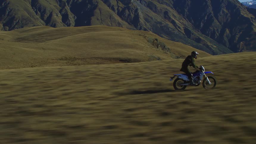 Motorcyclist stands on motorcycle while riding rapidly across field with view of mountain range